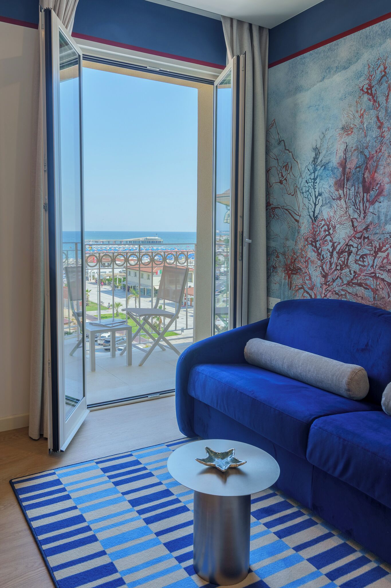 Une pièce avec un canapé bleu, une table ronde et une vue sur une plage et des bâtiments, visible à travers une porte vitrée.