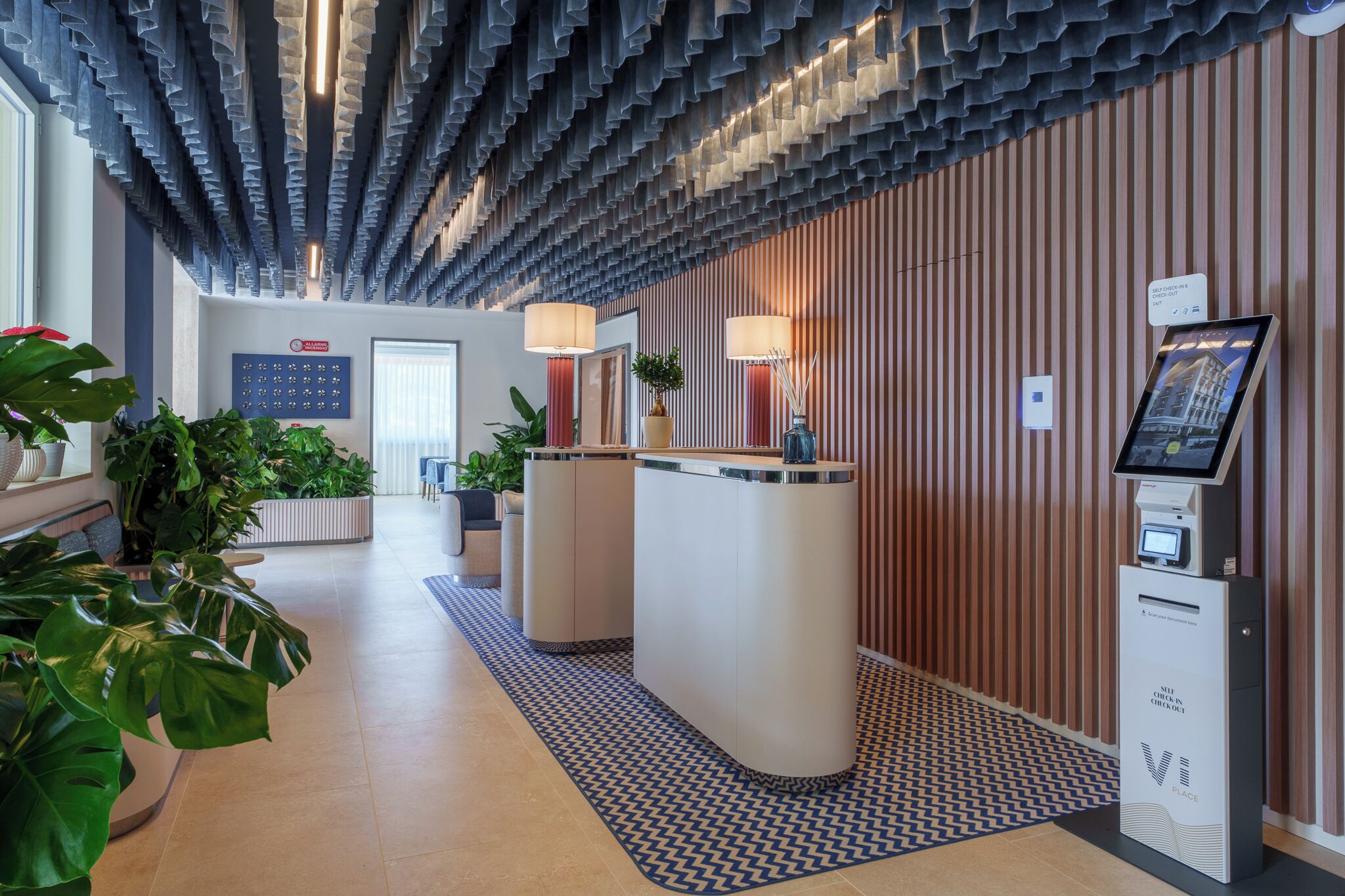 A hotel reception area with a modern design, featuring a check-in kiosk, a reception desk, and a patterned floor.