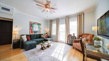 A living room with a sectional sofa, a coffee table, a glass table, a television, and a ceiling fan.