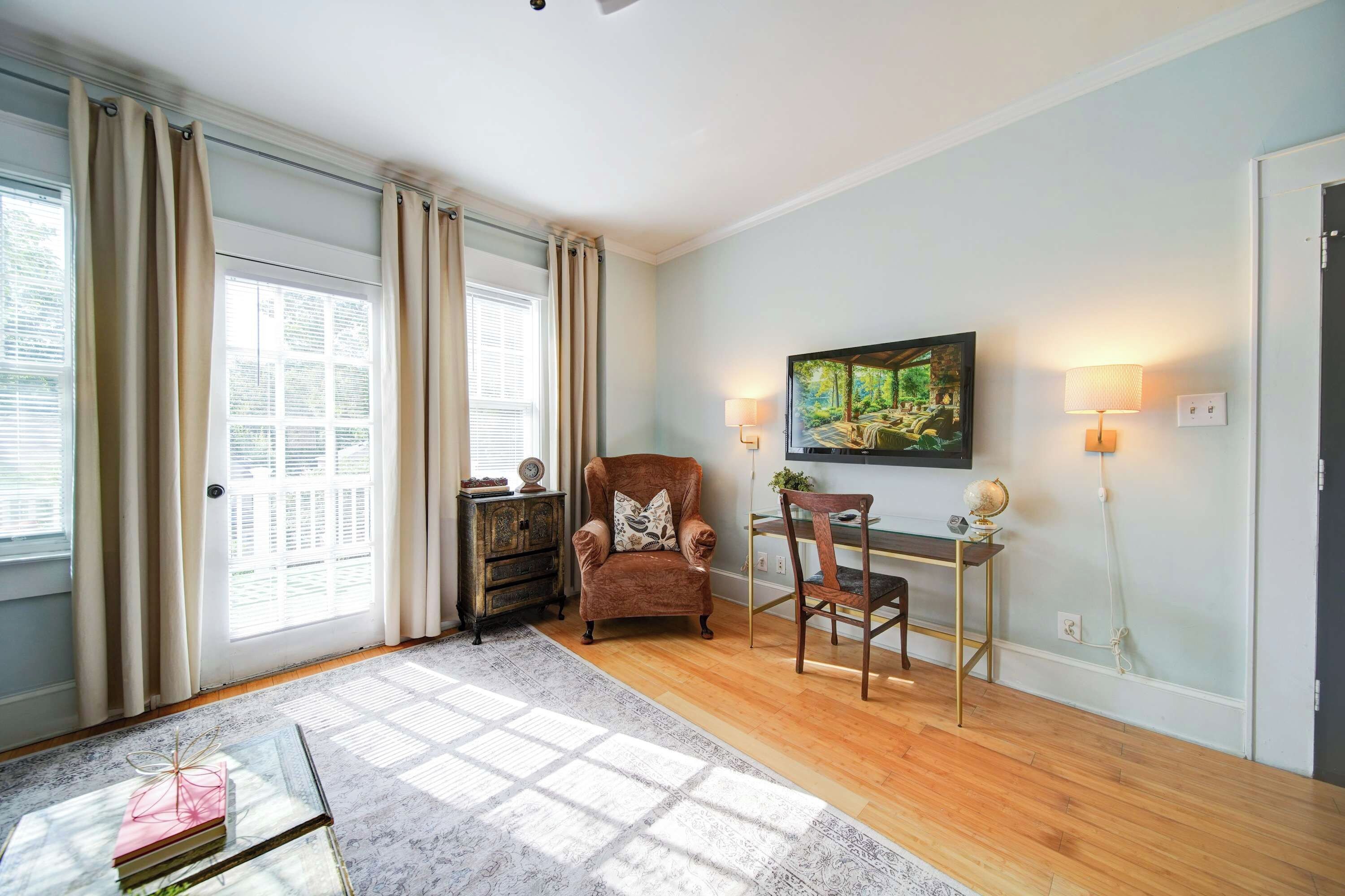 A room with a wooden floor, a brown armchair, a glass coffee table, a painting on the wall, and a desk with a chair.