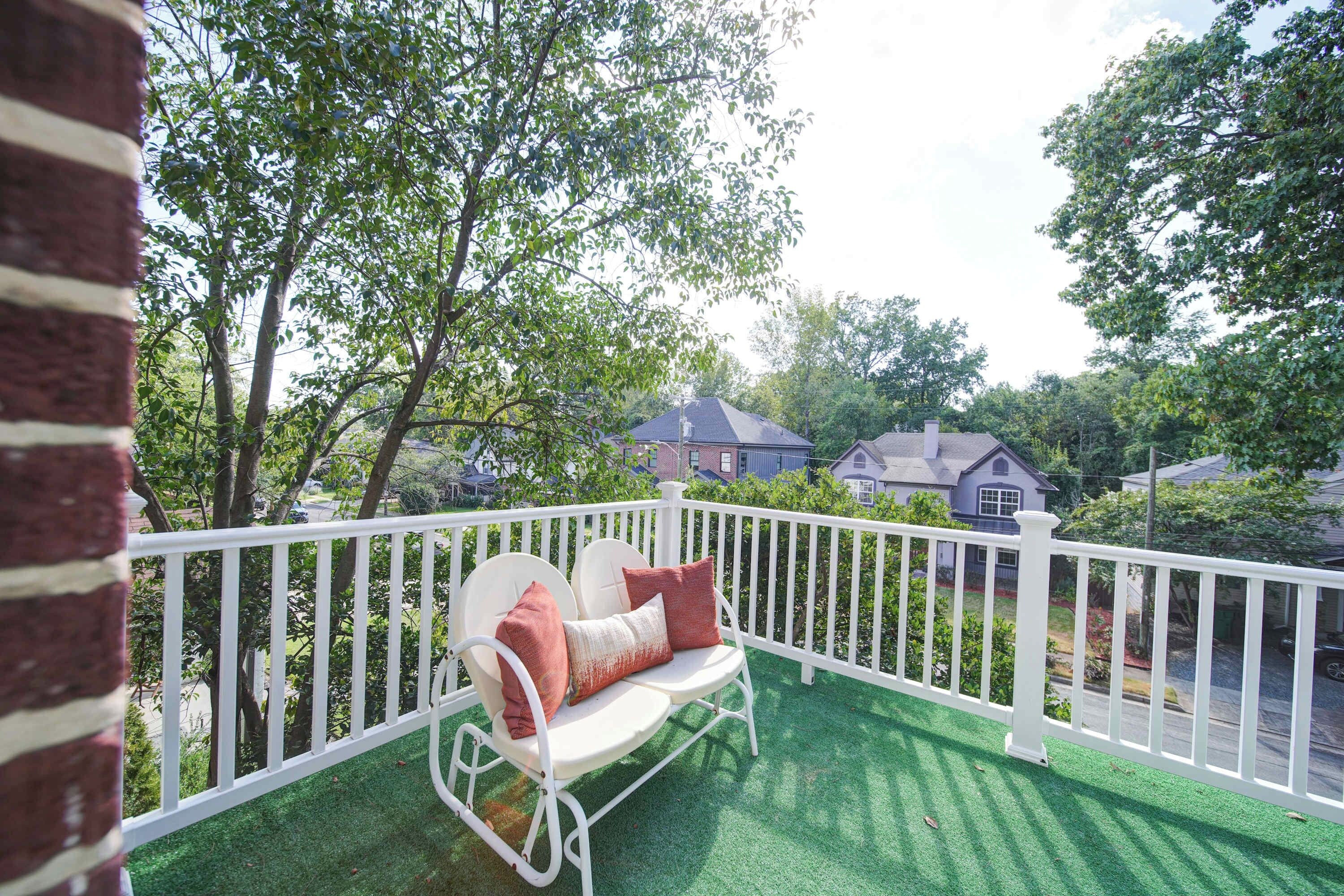 A balcony with a white railing, a white outdoor chair with cushions, and a view of houses in the background.