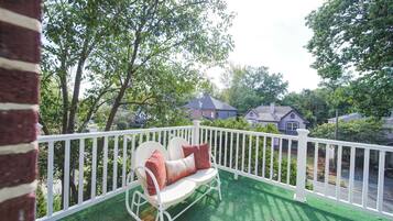 A balcony with a white railing, a white outdoor chair with cushions, and a view of houses in the background.