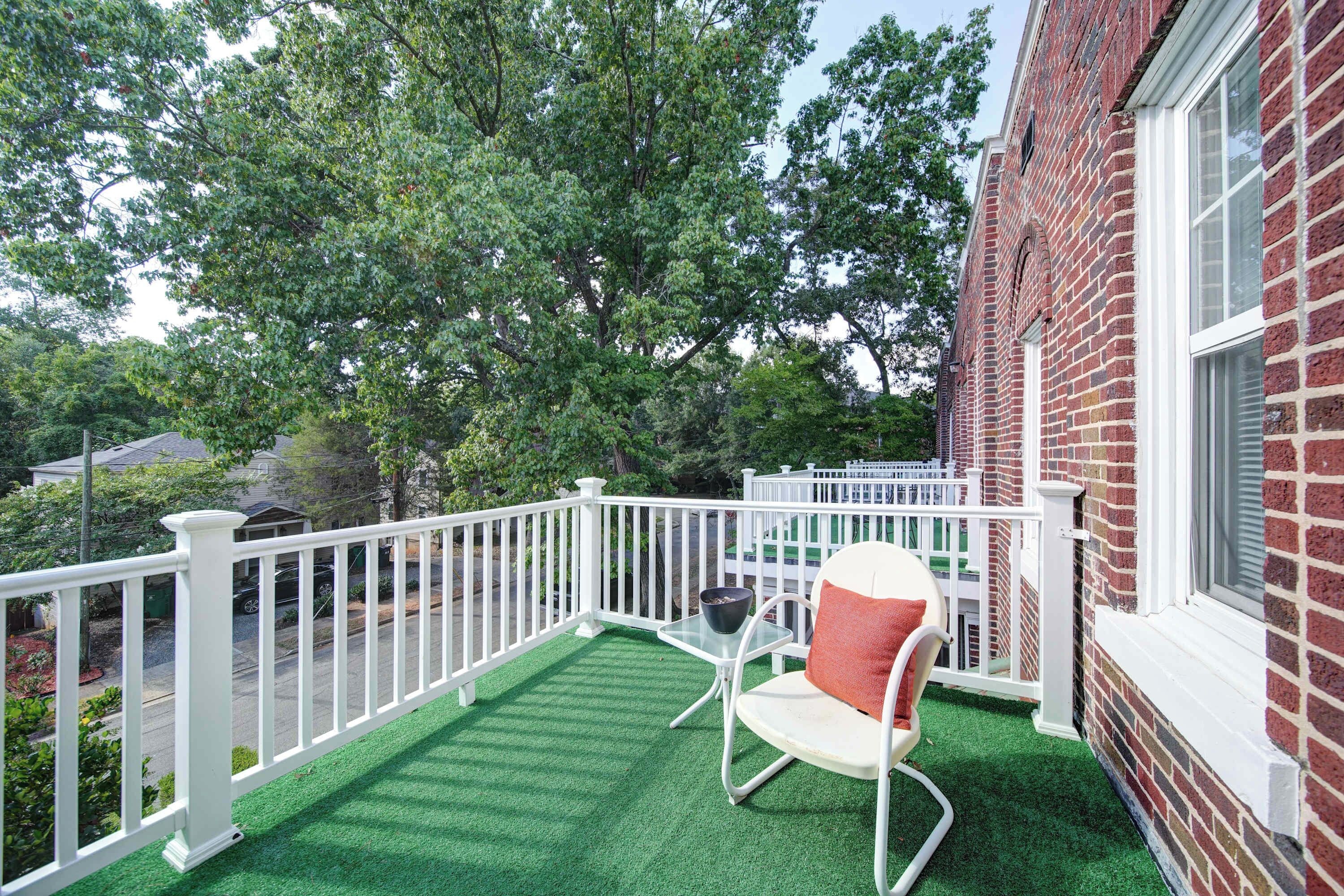 Un balcon avec une balustrade blanche, une seule chaise blanche et, au fond, un immeuble en briques.