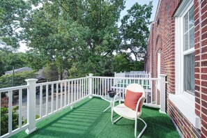 Un balcon avec une balustrade blanche, une seule chaise blanche et, au fond, un immeuble en briques.