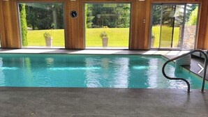 An indoor swimming pool with a view of a grassy area and trees through large windows. - Cozy apartment with pool and golf nearby (Megève)