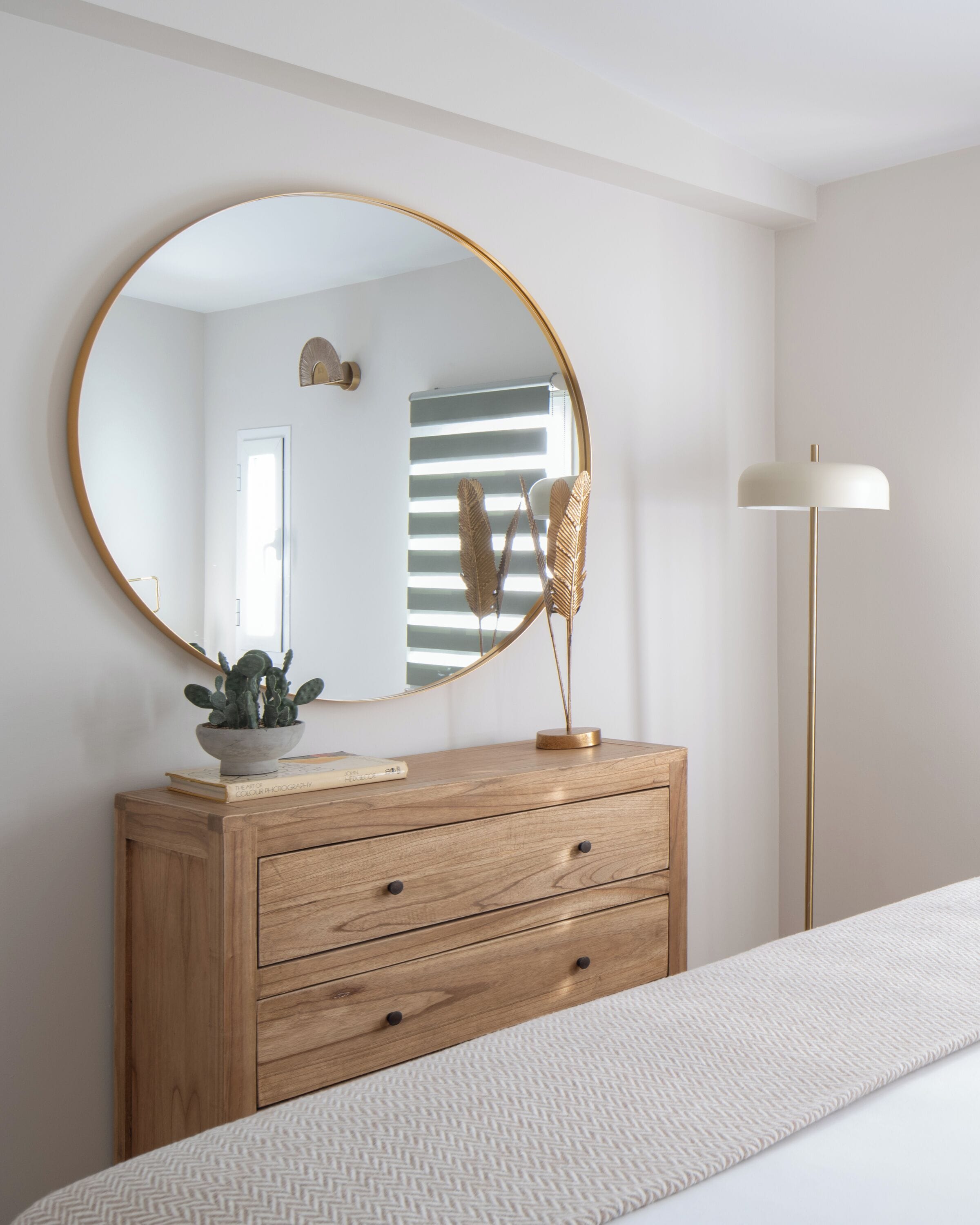 Een slaapkamer met een houten dressoir, een ronde spiegel, een vloerlamp en een bed met een bedovertrek met dessin.