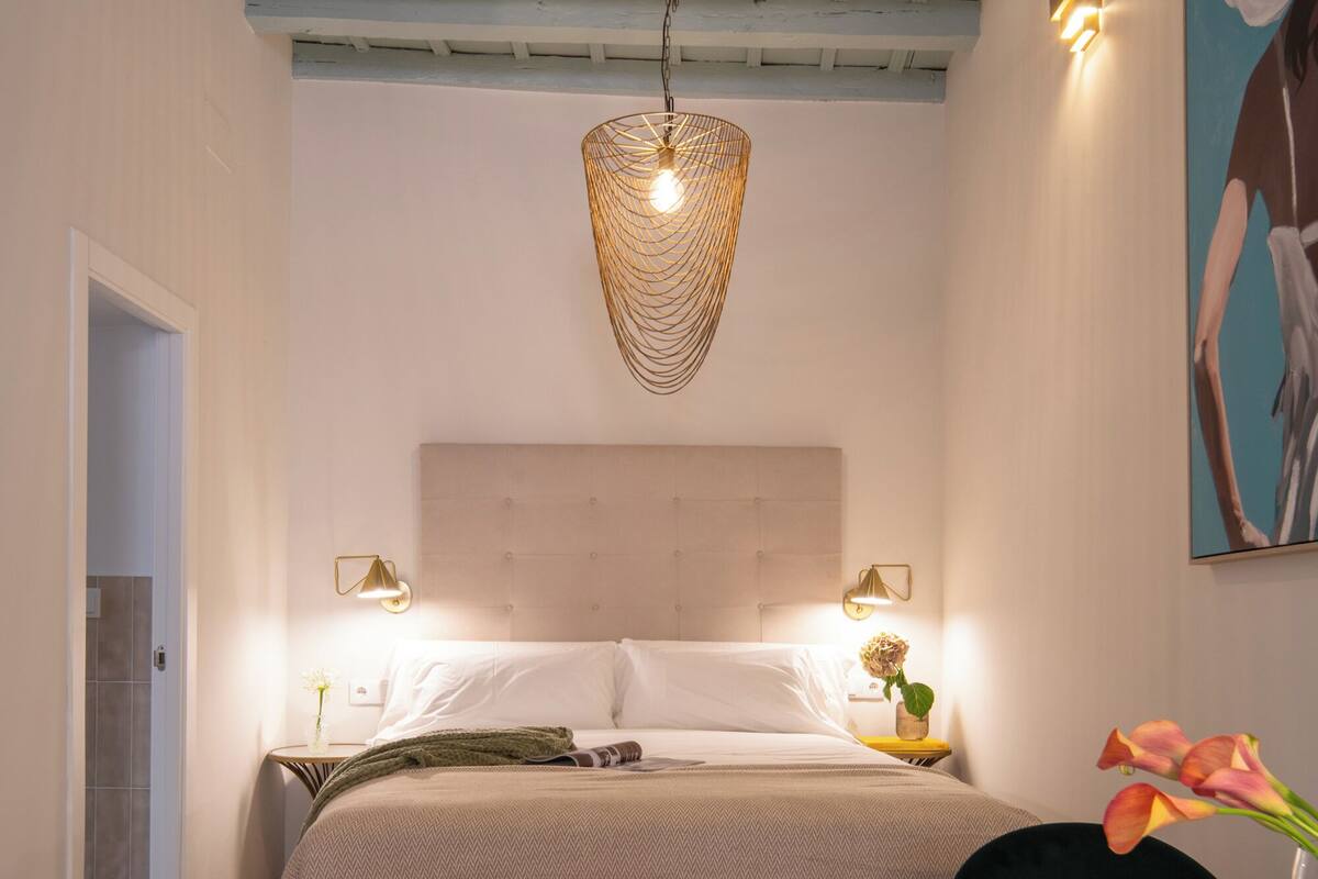 Un dormitorio con cama, mesitas de noche, una lámpara colgante y un cuadro en la pared.