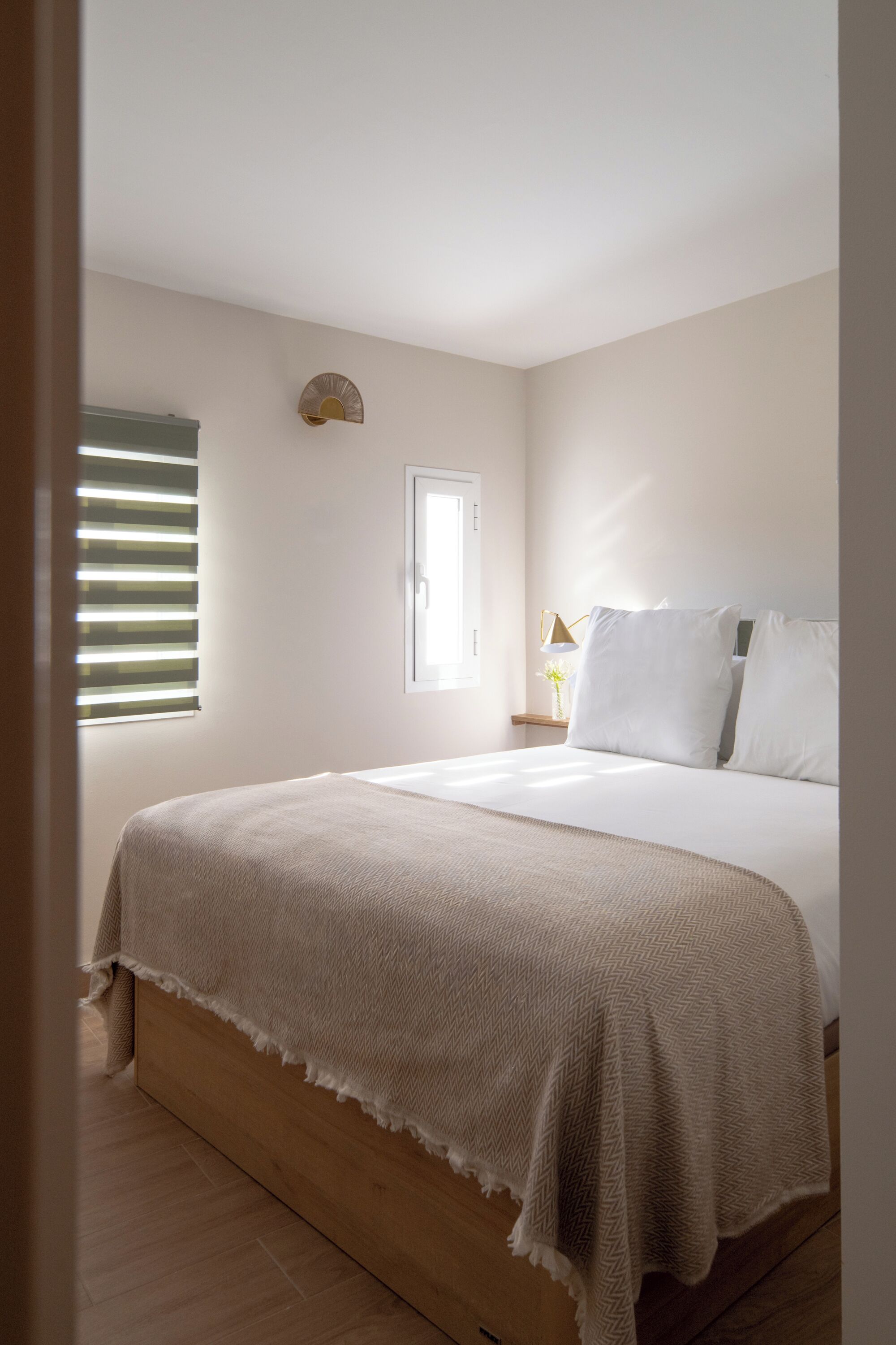 Un dormitorio con cama, ventana con persianas, mesita de noche con lámpara y una lámpara de pared.
