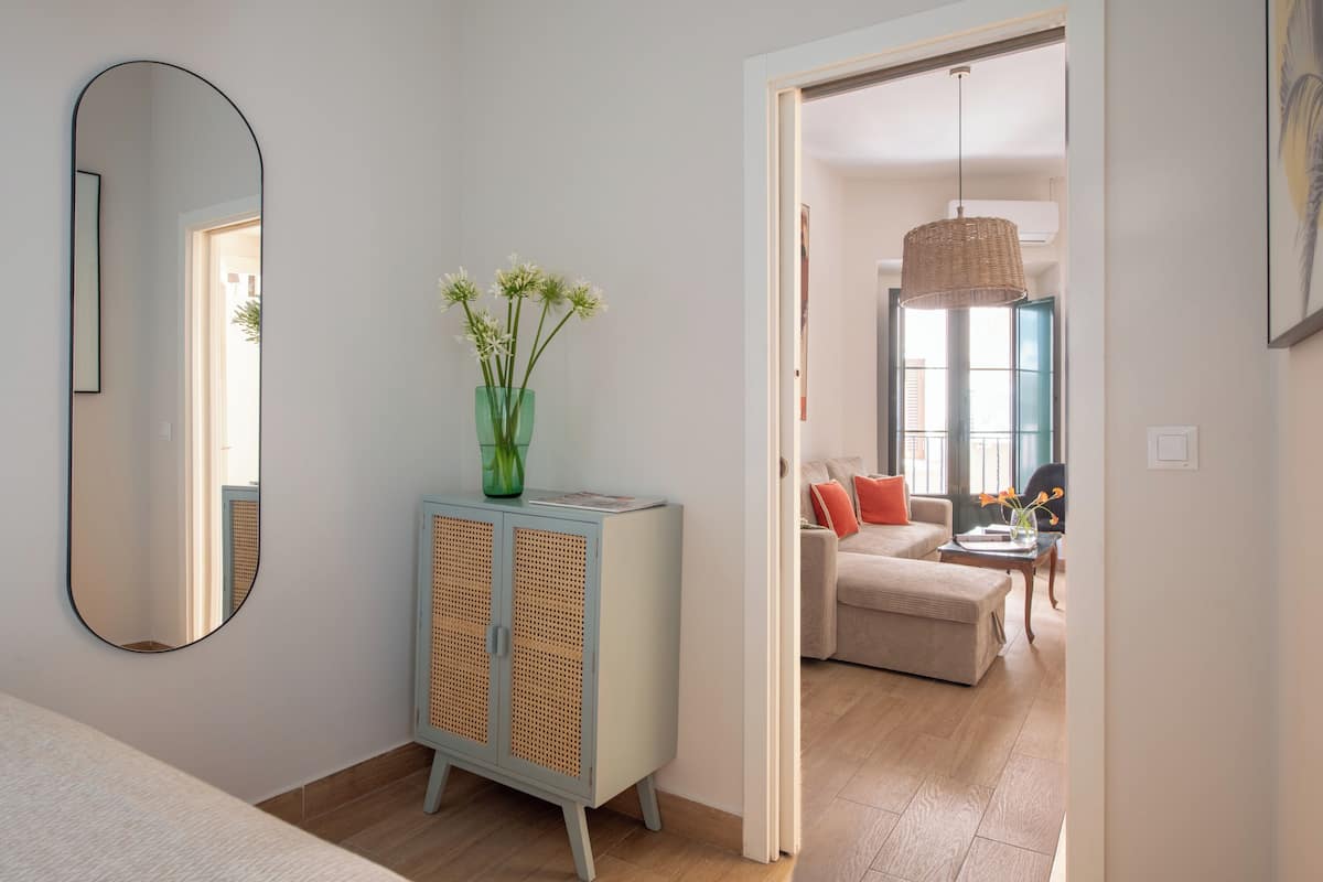 Una sala de estar moderna con un sofá, una mesa de centro y una mesa de comedor. Se observa un mueble con espejo y un florero con flores. La habitación tiene piso de madera y un ventanal grande.