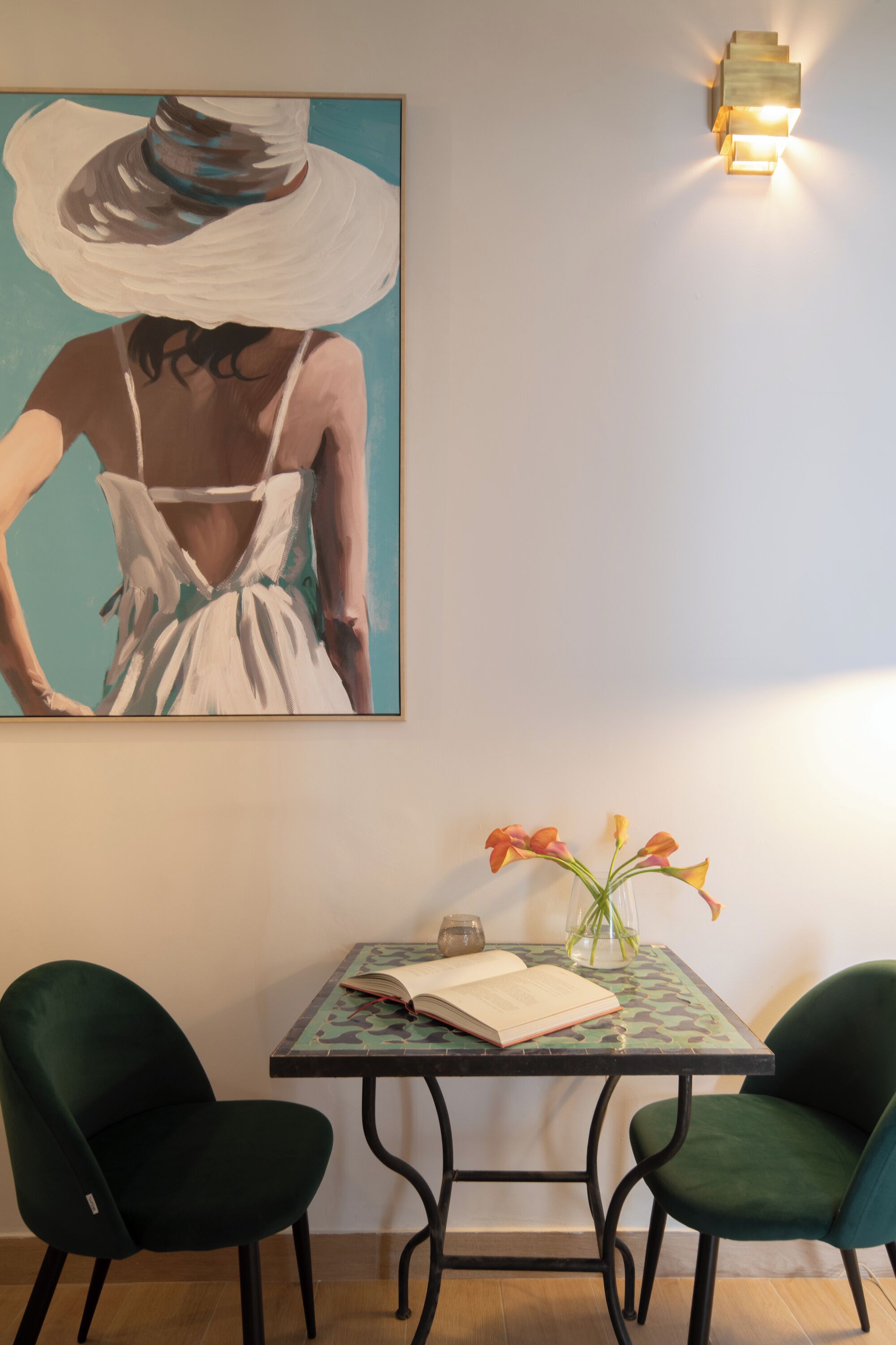 Une pièce dans laquelle on peut voir un tableau représentant une femme portant un chapeau, une table avec un livre et des fleurs, ainsi que deux chaises.