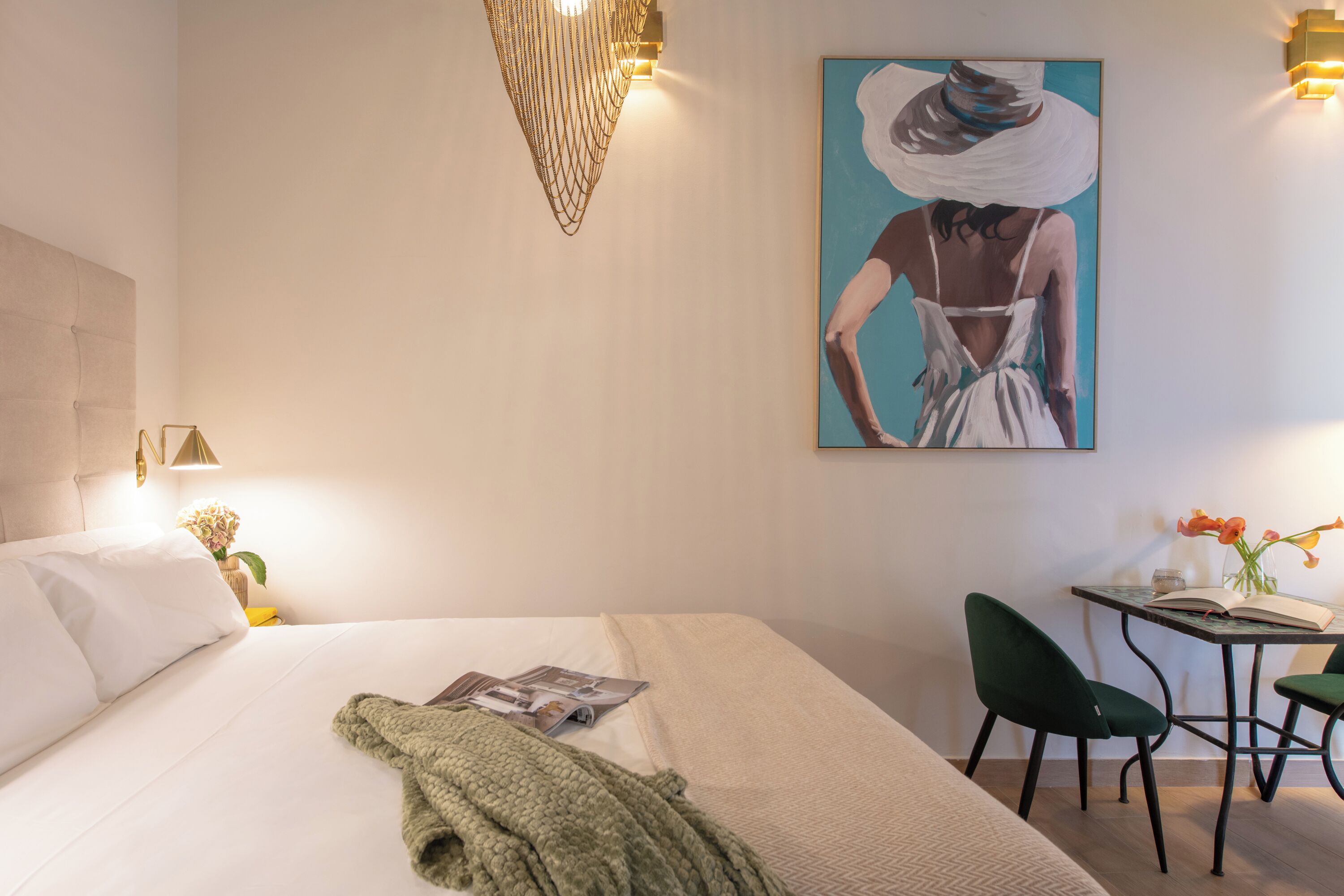 A hotel room with a bed, a desk with two chairs, a painting on the wall, and a vase with flowers on the desk.