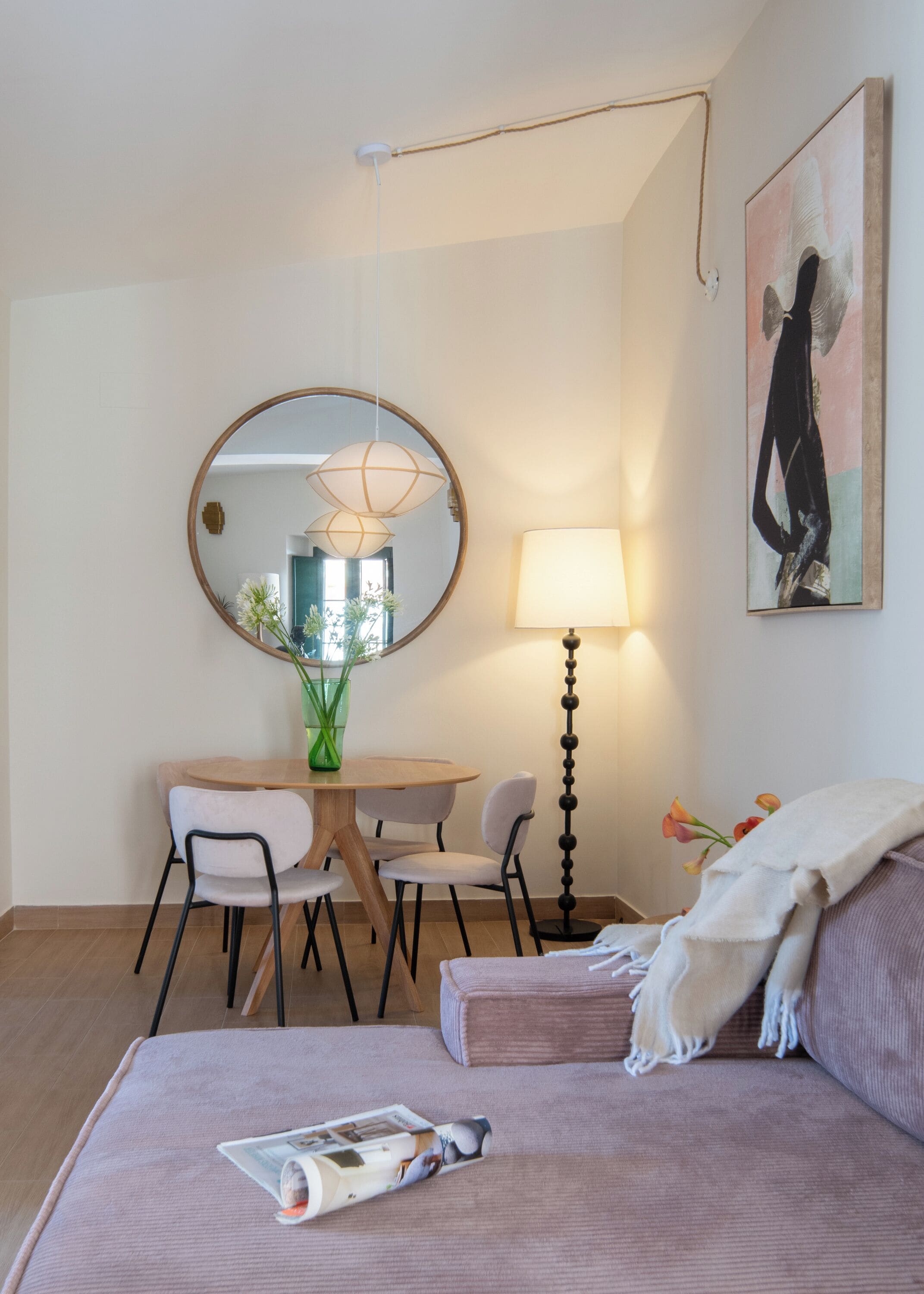 Una habitación de hotel moderna con un espejo redondo, una mesa de comedor con sillas, una lámpara de pie, una cama con una revista y un cuadro en la pared.
