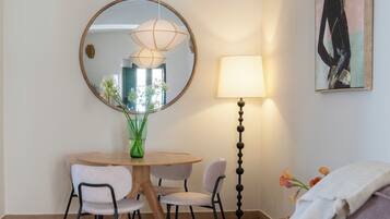 Una habitación de hotel moderna con un espejo redondo, una mesa de comedor con sillas, una lámpara de pie, una cama con una revista y un cuadro en la pared.