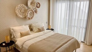 A bedroom with a large bed, a wall-mounted decorative piece, a bedside table with a lamp, and a window with sheer curtains. - Almadies Peaceful Oasis | 24/7 Security |Casa Molo (Dakar)