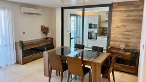 A modern dining area with a glass-top table and wooden chairs, a wooden cabinet with a plant, and a kitchen visible through a glass door. - Almadies Peaceful Oasis | 24/7 Security |Casa Molo (Dakar)
