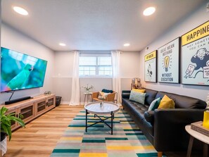 A modern living room with a flat-screen TV, a round coffee table, a sofa, and a colorful rug. - Playful, walkable, and comfortable, just steps from Mass Street. (Lawrence)