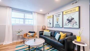 A modern living room with a black leather sofa, a round coffee table, and a colorful striped rug. There are three motivational posters on the wall and a window with white curtains.