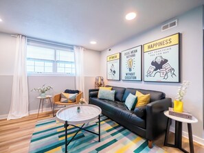 A modern living room with a black leather sofa, a round coffee table, and a colorful striped rug. There are three motivational posters on the wall and a window with white curtains. - Playful, walkable, and comfortable, just steps from Mass Street. (Lawrence)