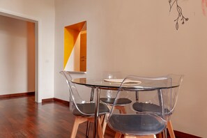 A dining area with a glass table and four chairs, a wooden floor, and a white wall with a geometric decoration.