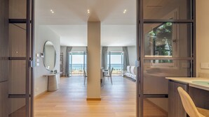 A modern hotel room with a wooden floor, a large mirror, a dining area with a view, and a kitchen area.