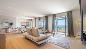 A modern living room with a sofa, dining area, and large windows offering a view of the sea.