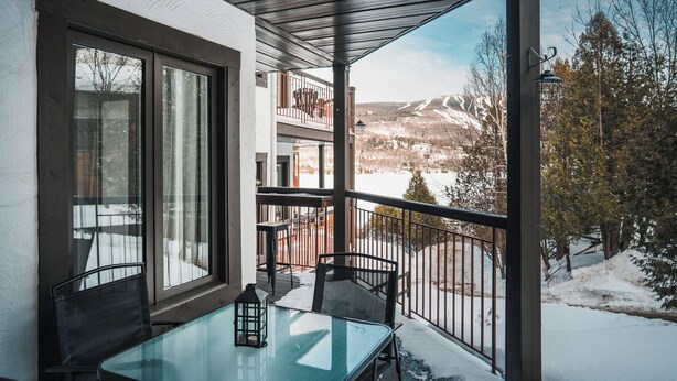 A balcony with a glass table and chairs, overlooking a snowy landscape with mountains.