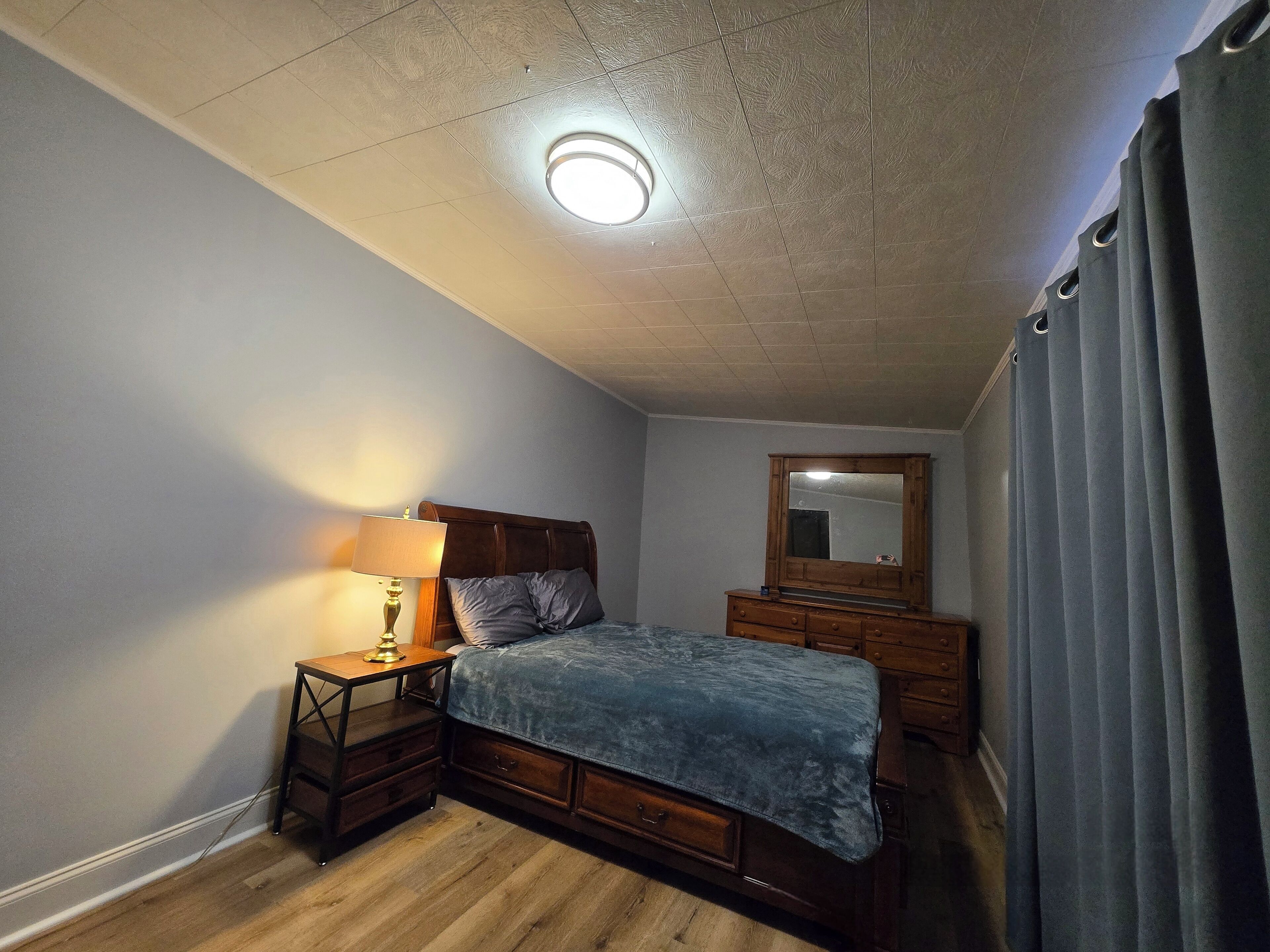 A bedroom with a wooden bed, a nightstand with a lamp, a dresser, a mirror, and a window with curtains.