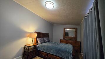 A bedroom with a wooden bed, a nightstand with a lamp, a dresser, a mirror, and a window with curtains.