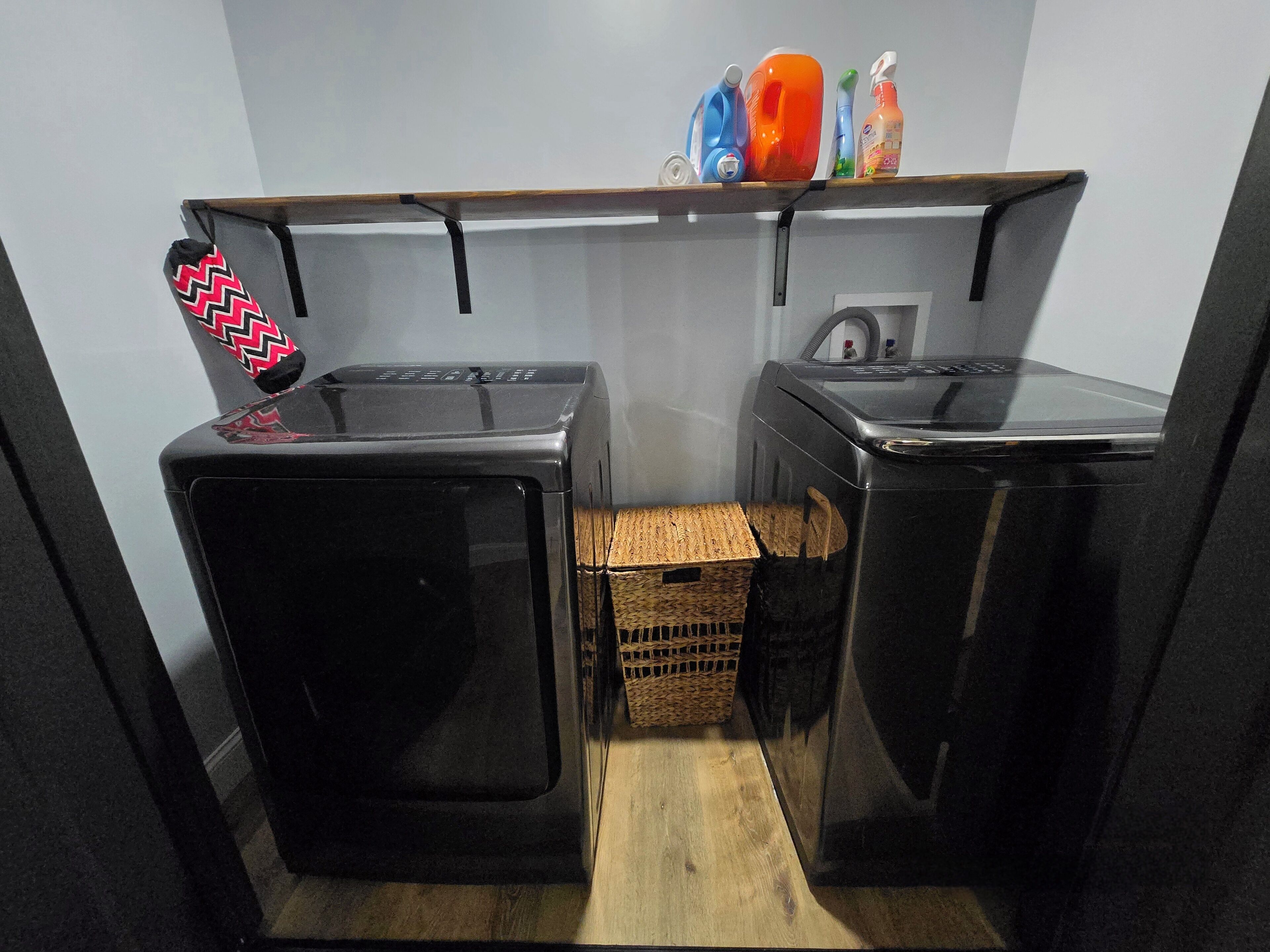 A compact laundry room with two black washing machines, a wooden shelf with cleaning supplies, and a wicker basket.