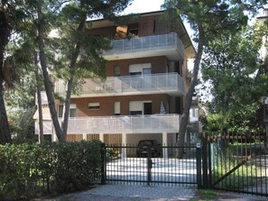 Um edifício residencial com vários andares, varandas, portão fechado e árvores ao redor.