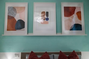 Tres obras de arte abstractas enmarcadas sobre una pared verde azulada, encima de una cama con almohadones rojos.