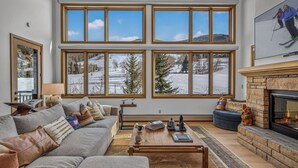Ein Wohnzimmer mit großem Fenster mit Blick auf eine verschneite Landschaft, einem Kamin und einer gemütlichen Sitzecke.