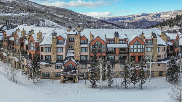 Ein mehrstöckiges Hotel mit Steinen und Holzfassade, eingebettet in eine verschneite Berglandschaft mit Skipisten im Hintergrund.