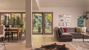 Ein modernes Wohnzimmer mit einem Ecksofa, einem Esstisch mit Stühlen und einem großen Fenster mit Blick auf Bäume.