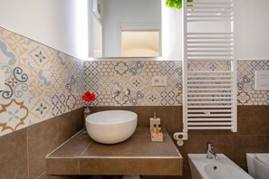 A bathroom with a large white sink, a mirror, a towel rack, and patterned tiles. - GuestHost - Charming room of about 40 square meters, ideal for two people, located on the ground floor of a historic building (there is a small step at the entrance). Furnished with taste and attention to detail, it offers a warm and suggestive atmospher (Bari)