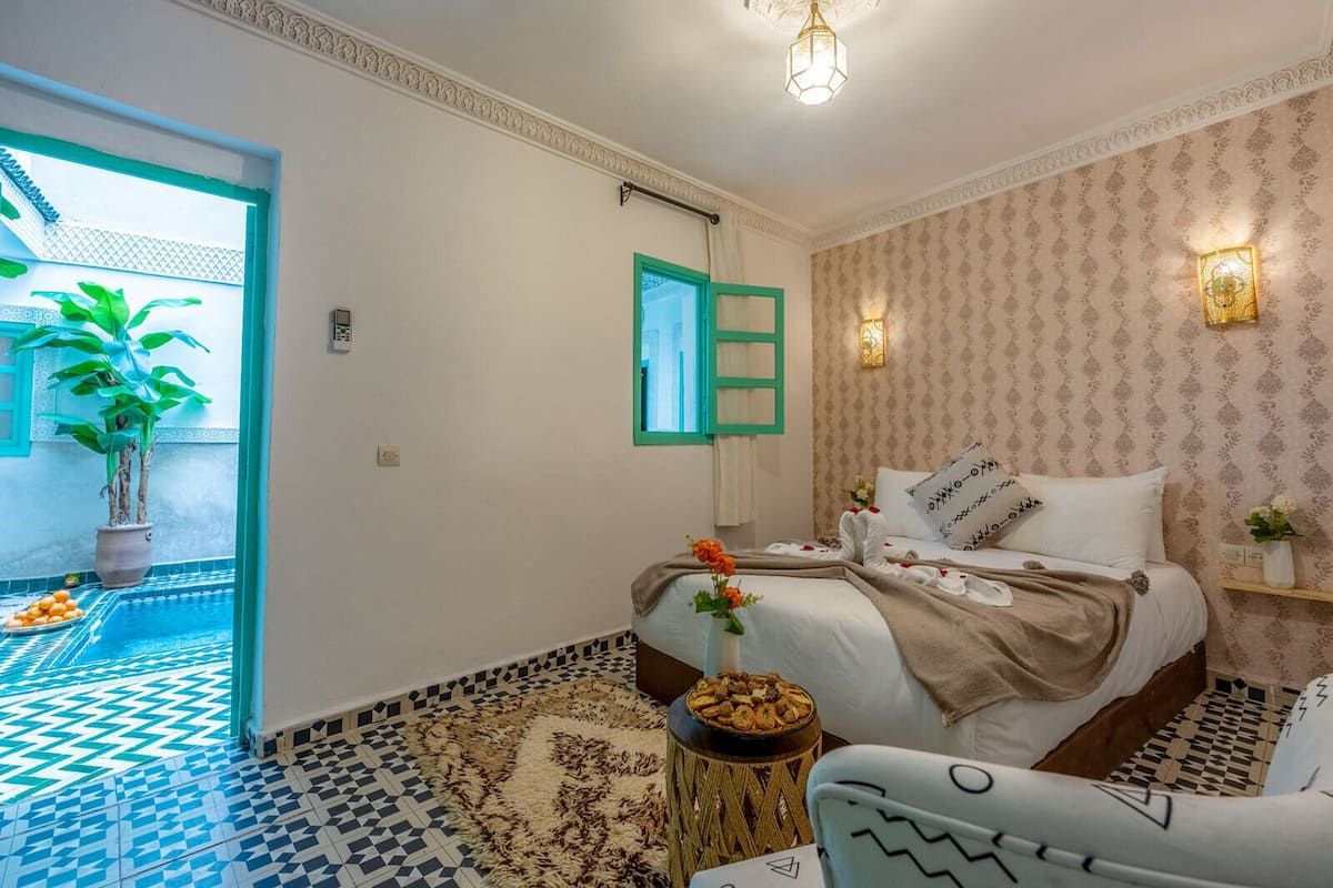 A bedroom with a bed, a bedside table, a chair, a potted plant, and a view of the outside.