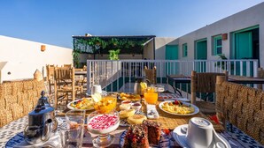 An outdoor dining area with wicker furniture, a variety of breakfast dishes, and a view of a building with green shutters. - Riad JABADOR De Marrakech (Marrakech)