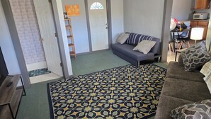 Una sala de estar con un sofá gris, un tapete estampado, una escalera de madera y una puerta que da a otra habitación.