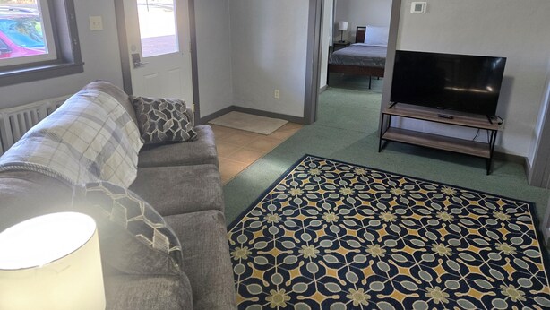 Una sala de estar con un sofá, un tapete estampado, un televisor sobre un mueble bajo y una puerta que da a un dormitorio.