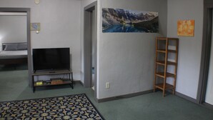 Una sala de estar con un televisor, una escalera de madera, un cuadro de un paisaje montañoso y una alfombra estampada.