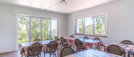 A dining area with multiple tables and chairs, large windows with a view of greenery, and a ceiling with a visible light fixture.