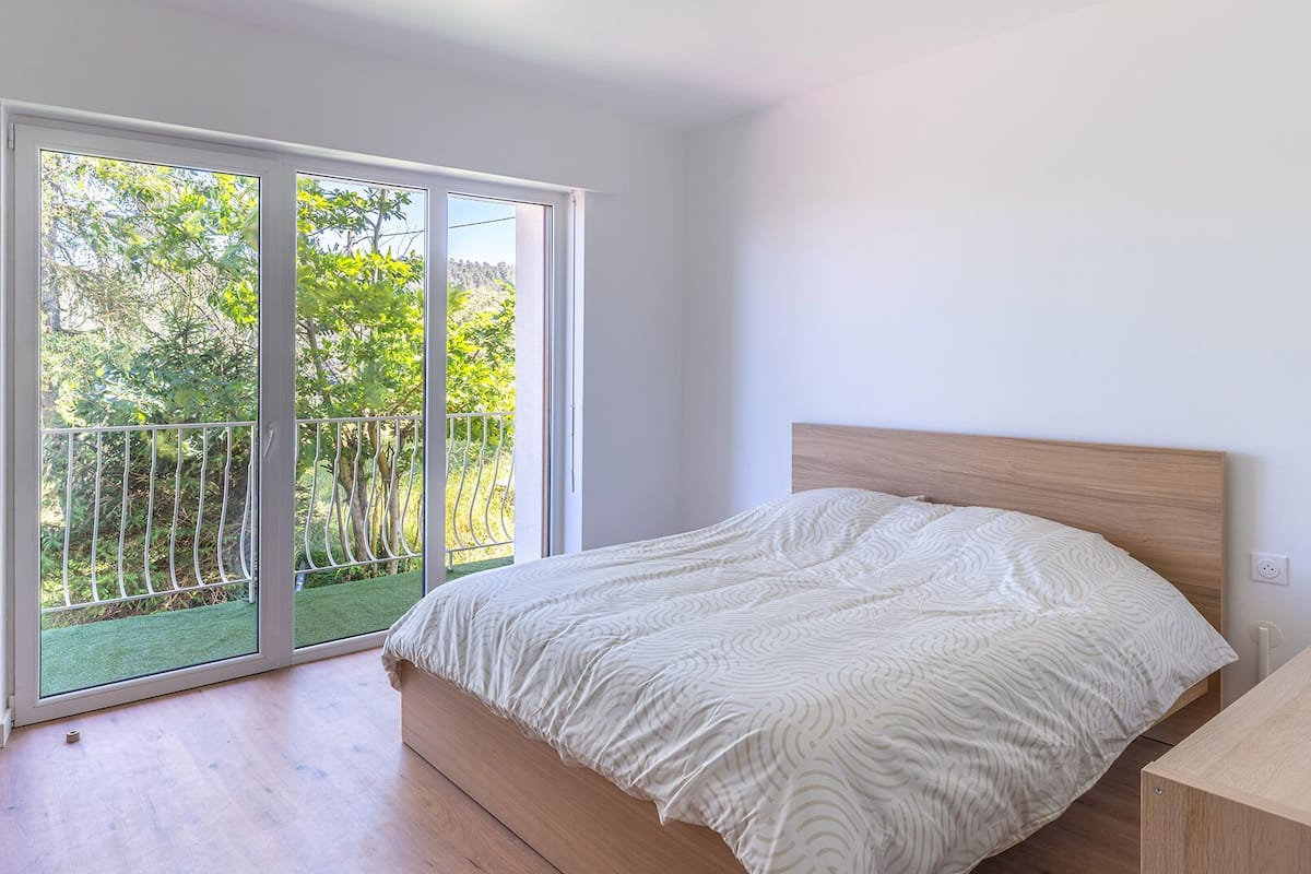 Una camera da letto con un letto, una testiera in legno e una porta scorrevole in vetro che dà su un balcone con vista sugli alberi.