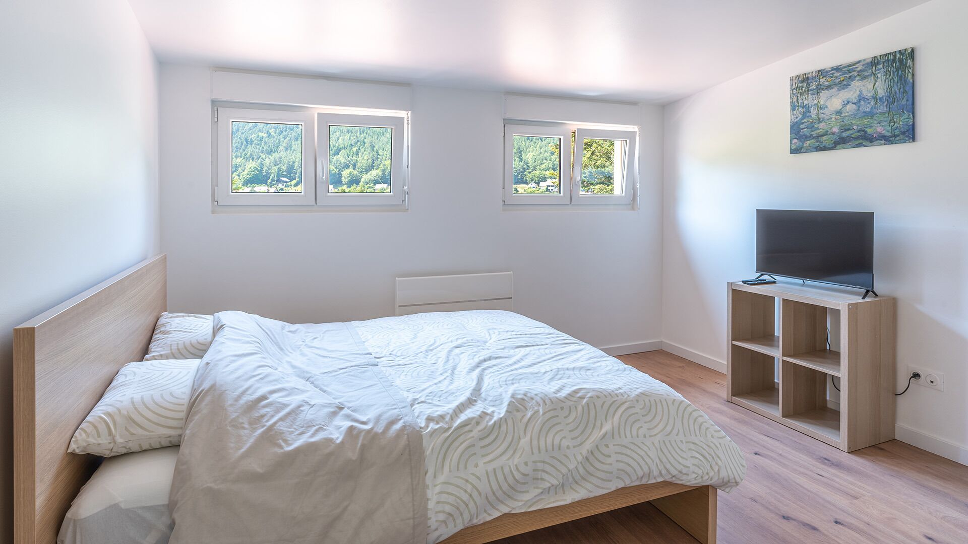 Un dormitorio con cama, televisor sobre un mueble de madera y tres ventanas con vistas a zonas verdes.