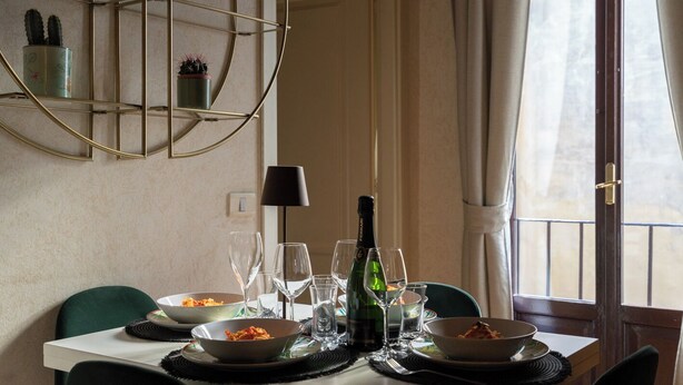 A dining table set with plates, glasses, and a bottle of wine, with chairs and a decorative metal shelf in the background. - Violetta's Home (Firenze)