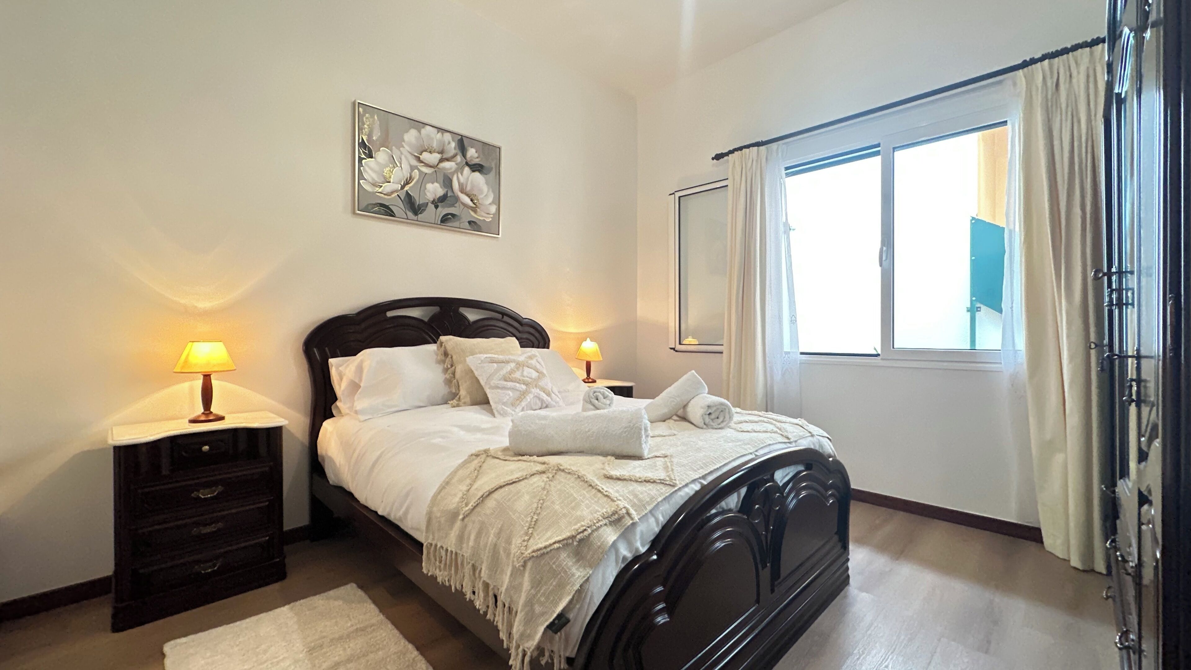 Una cama bien hecha con ropa de cama blanca, dos lámparas de noche, un cabecero de madera oscura, una manta blanca y un cuadro de flores en la pared.