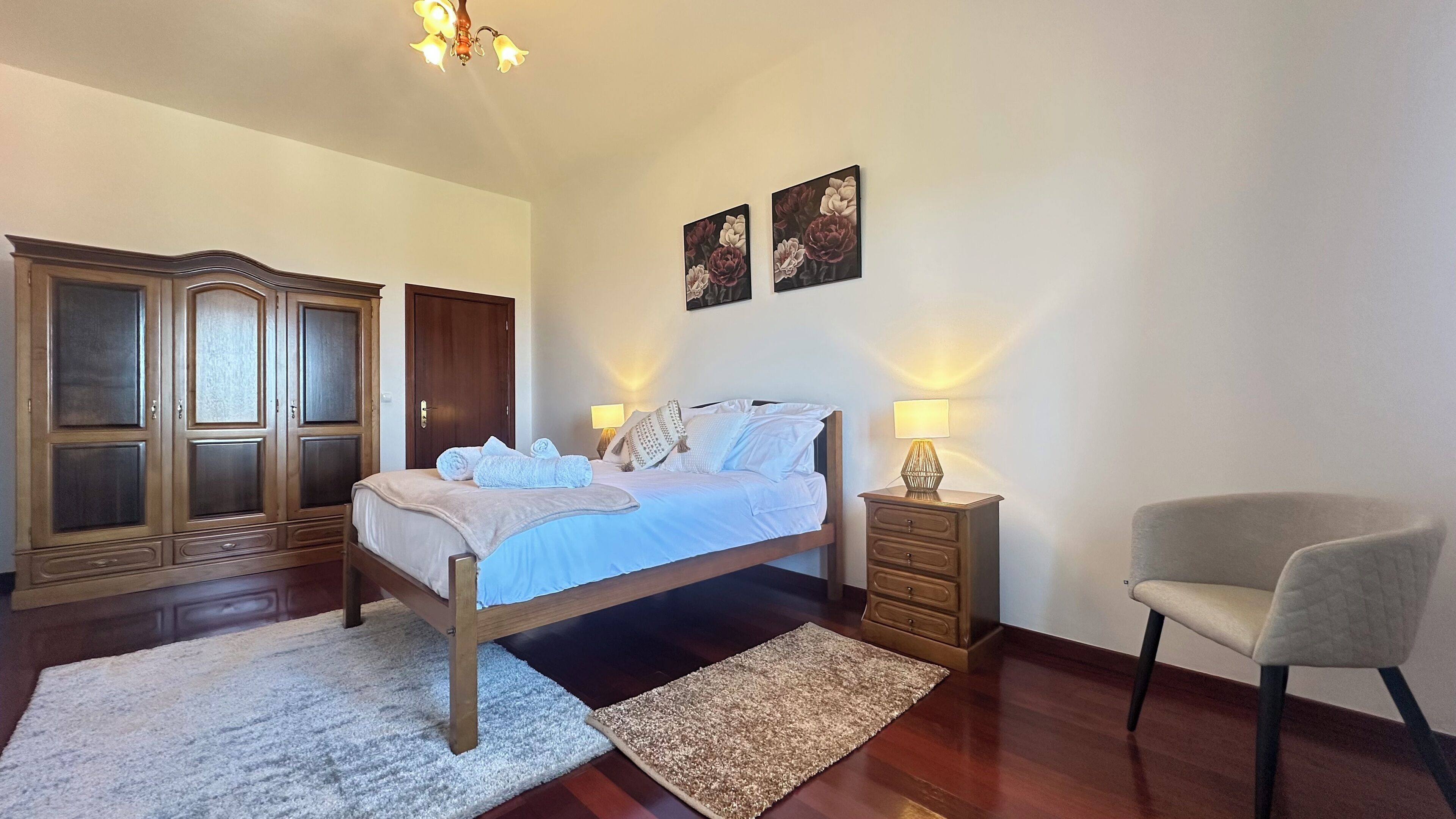 A bedroom with a bed, a nightstand, a chair, a wardrobe, and two wall-mounted lamps.