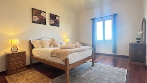 A bedroom with a wooden bed, two bedside lamps, a nightstand, a window with curtains, and two floral paintings on the wall. - Sea and Sun 4 You - Villa Jardim (Santana)