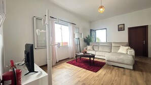 A spacious living room with a large sofa, a coffee table, and a TV. There is a red rug on the floor, and a fire extinguisher is visible on the left side. - Sea and Sun 4 You - Villa Jardim (Santana)