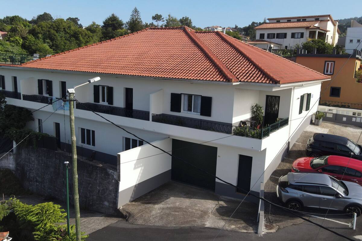 A two-story building with a red-tiled roof, a balcony with potted plants, and a parking area with several cars.