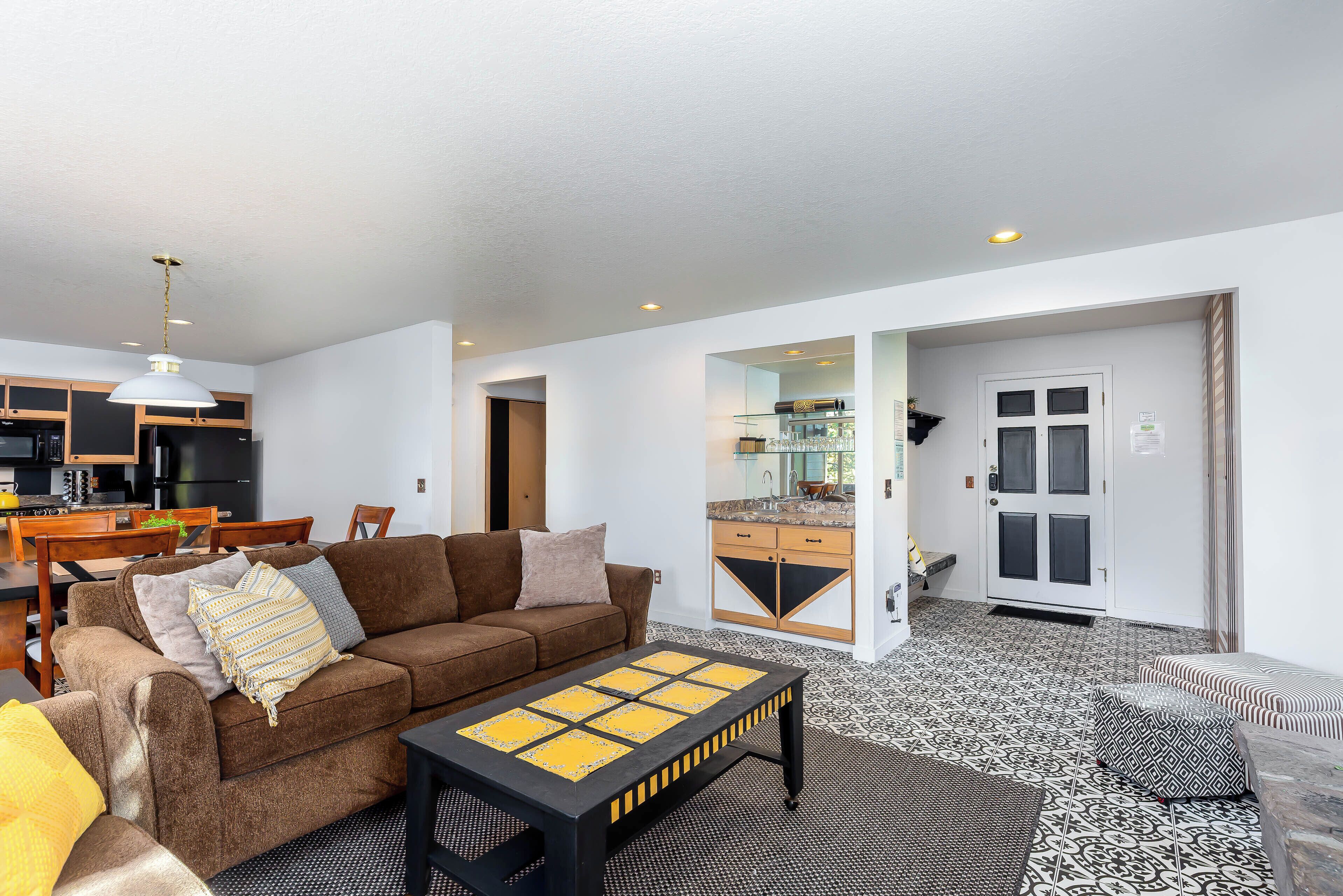 A spacious living area with a brown sofa, a coffee table, and a dining table with chairs. A kitchen with a black countertop and wooden cabinets is visible in the background.